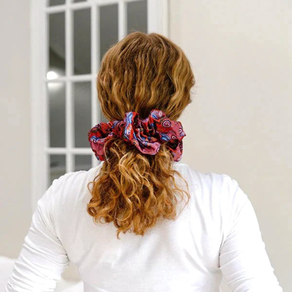 Person with red patterned scrunchie in hair, wearing a white shirt and beige pants, standing in a room.