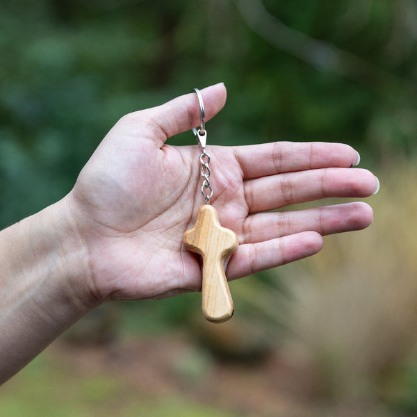 A small wooden cross keychain with a silver metal chain and split ring, held in a person's hand against a blurred outdoor green background.