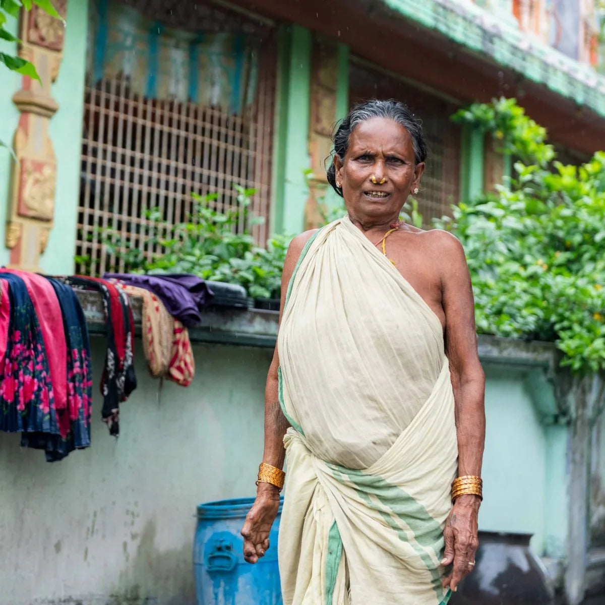Woman in a white saree standing outdoors with greenery and a building in the background