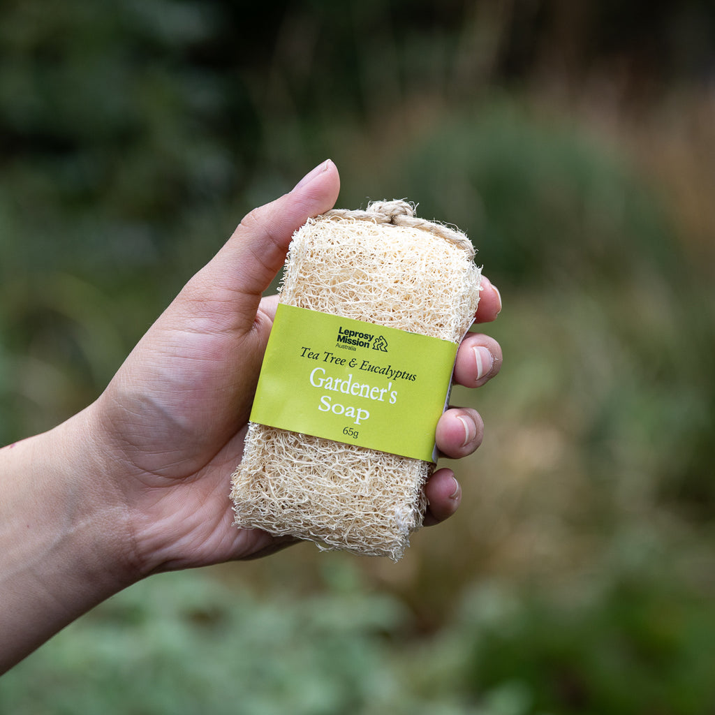 A hand holding a rectangular natural loofah soap bar wrapped with a lime green paper label that reads 'Lavender & Eucalyptus Gardeners Soap 6oz'. The background is a soft, blurred outdoor green garden setting.