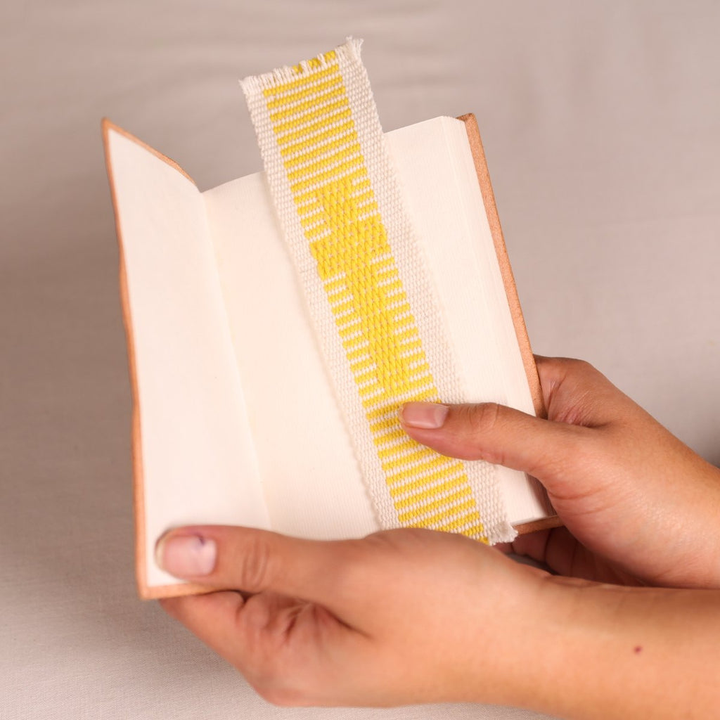 A close-up of a person holding an open book with a yellow and white woven fabric bookmark or page divider inserted between the pages.