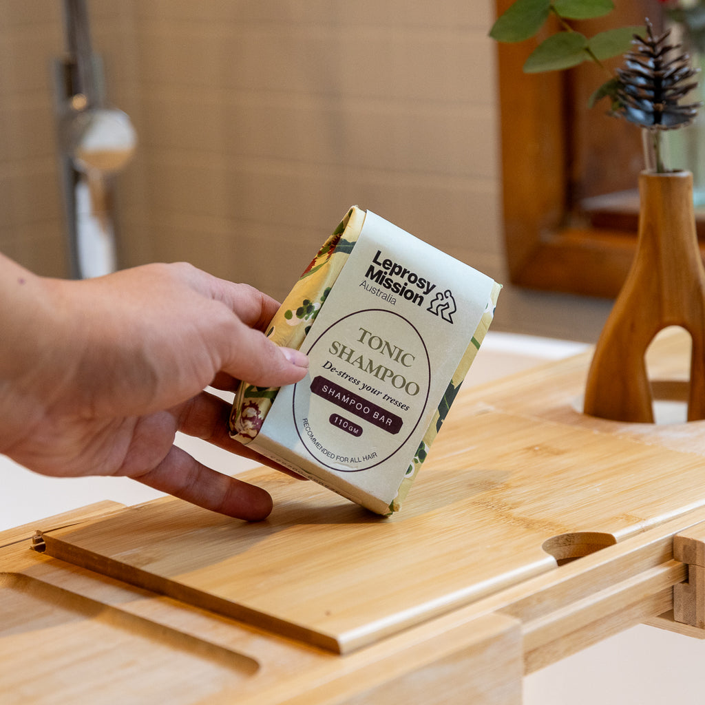 A hand holds a Leprosy Mission Tonic Shampoo bar over a wooden bath platform in a bathroom setting.