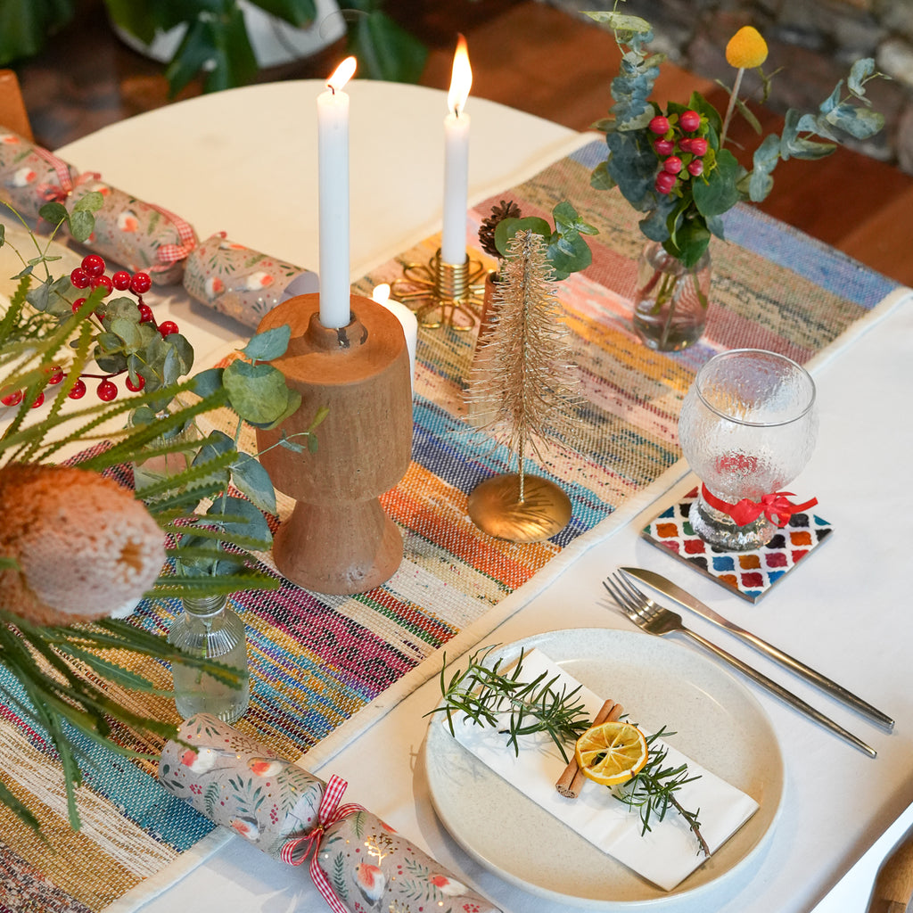 A styled holiday table setting with a colorful striped runner, white tablecloth, candle holders, wrapped crackers, a glass goblet, silverware, and dried botanical garnishes on plates.