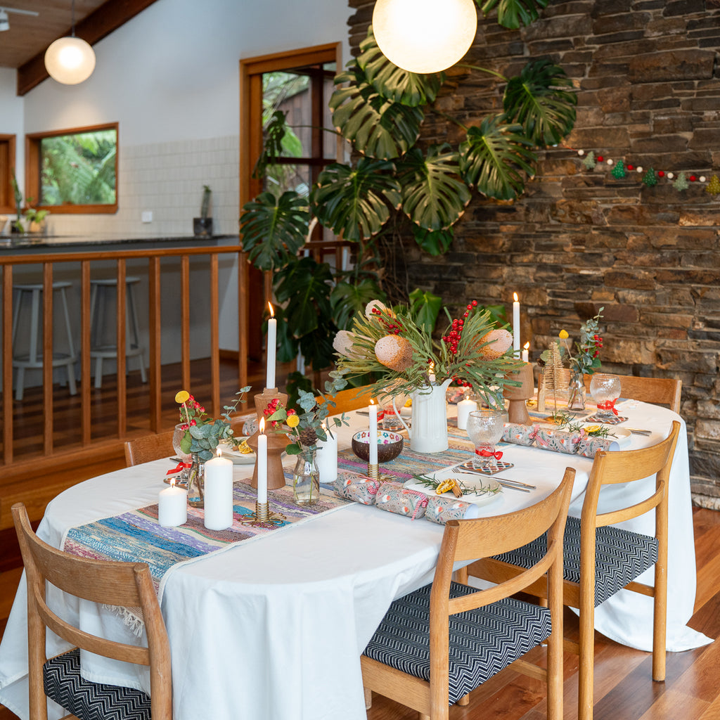 A styled dining table in a rustic indoor setting with a white tablecloth, floral centerpieces, lit candles, and wooden chairs. The room features a stone wall, large green tropical leaves, and pendant lights.