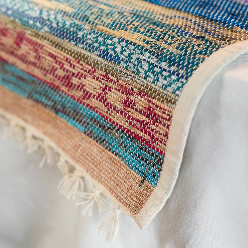 A close-up of a woven textile with a multicolor striped pattern in beige, blue, teal, pink, and brown, finished with a cream border and fringe tassels.