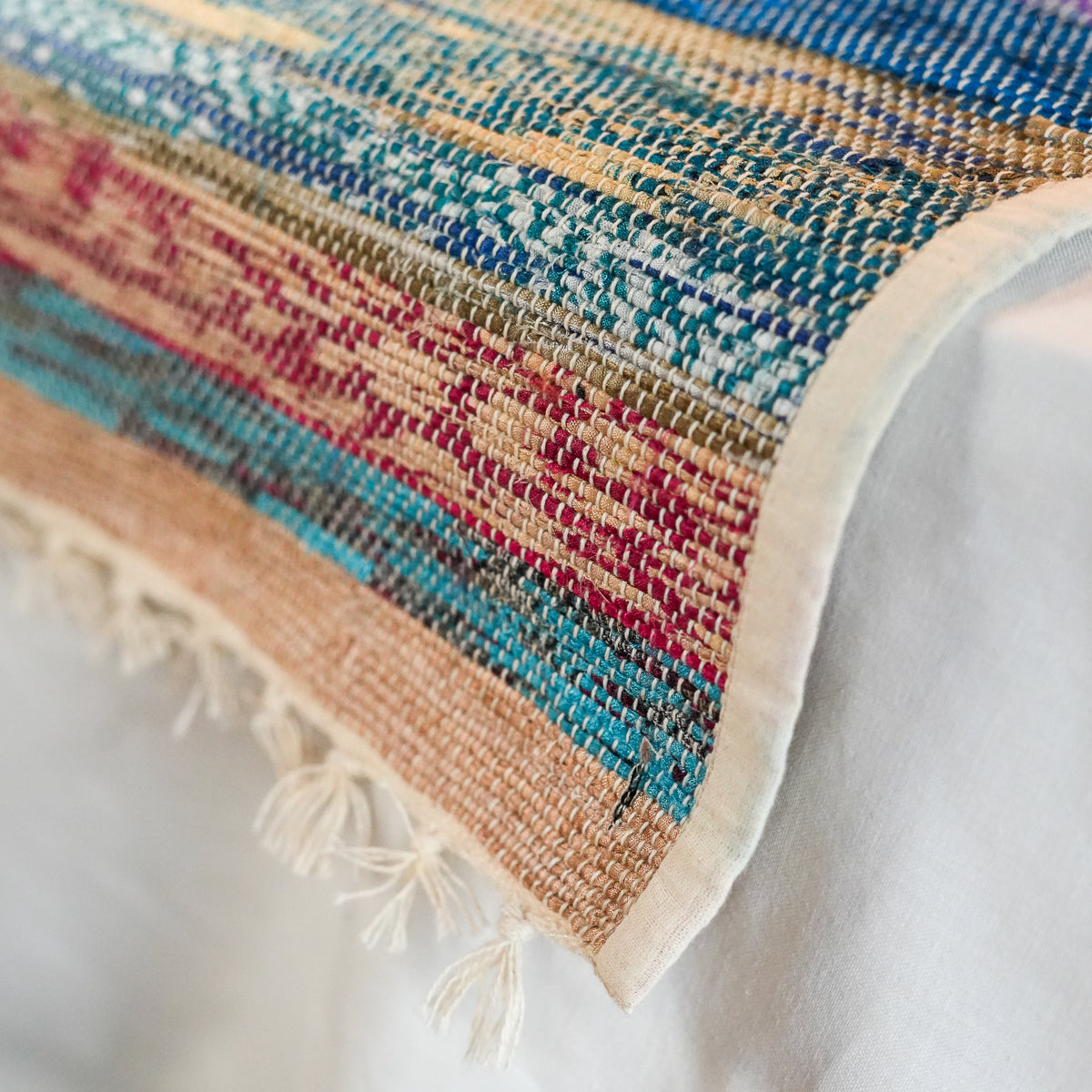 A close-up of a woven textile with a multicolor striped pattern in beige, blue, teal, pink, and brown, finished with a cream border and fringe tassels.