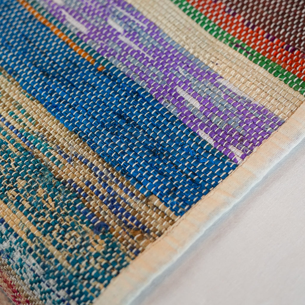 A close-up of a woven textile or rug showing a multicolored pattern with blue, purple, teal, beige, green, and red sections, finished with a cream border.