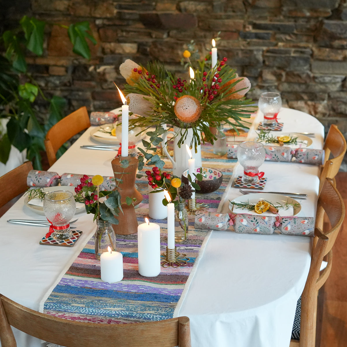 A styled round dining table set for a holiday or rustic celebration, featuring a colorful striped runner, white tablecloth, lit pillar candles, floral centerpieces, patterned napkins, and wrapped crackers in a warm indoor setting with a stone wall and plants.