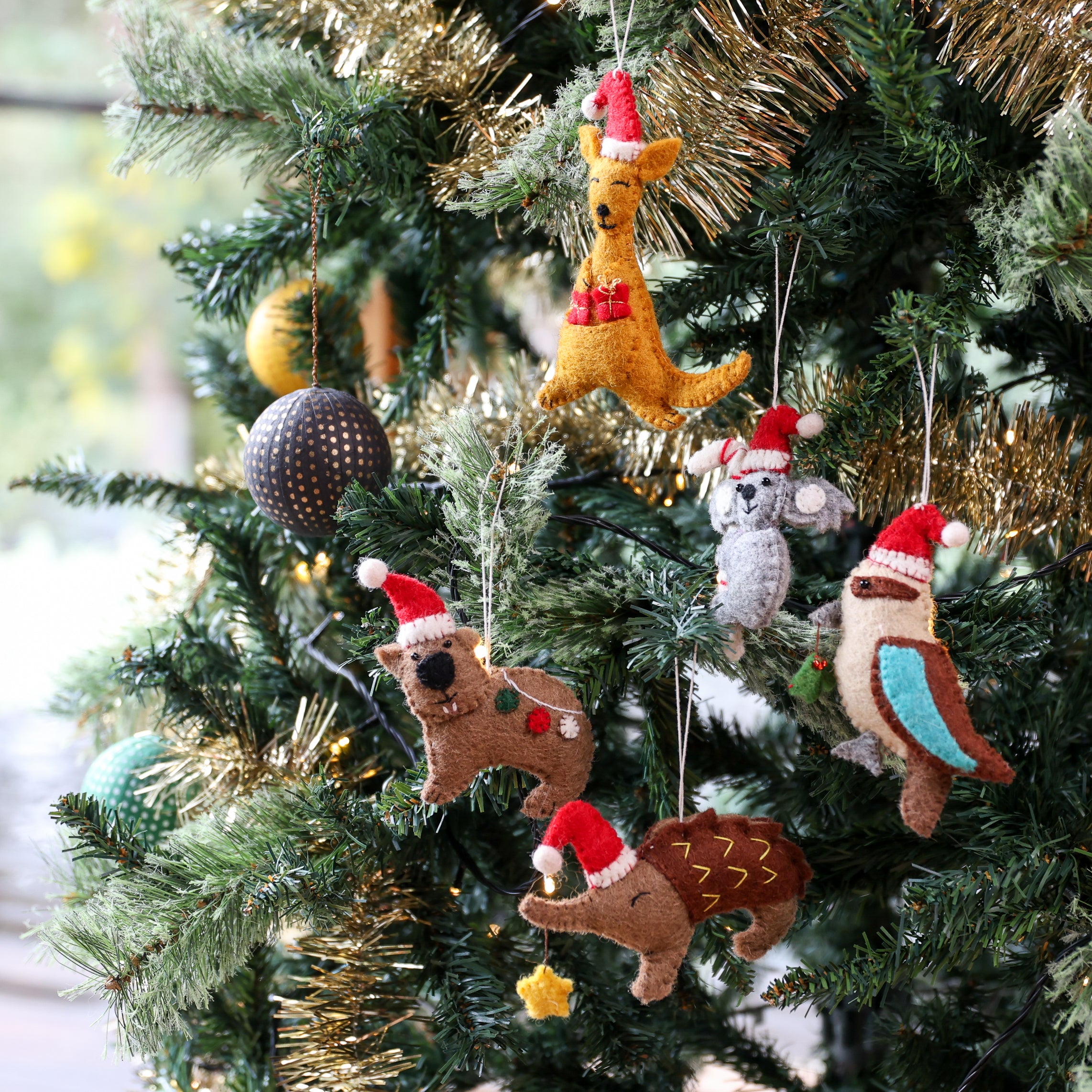 A collection of handmade felt animal Christmas ornaments hanging on a decorated green Christmas tree with gold tinsel. The felt animals include a kangaroo, a rabbit, a squirrel, a bird, and a dog, many wearing red Santa hats.