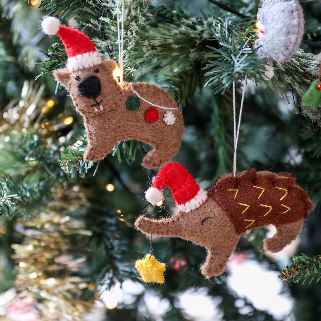 Two handmade felt Christmas tree ornaments shaped like a beaver and an elephant wearing Santa hats, hanging on a decorated Christmas tree with lights.