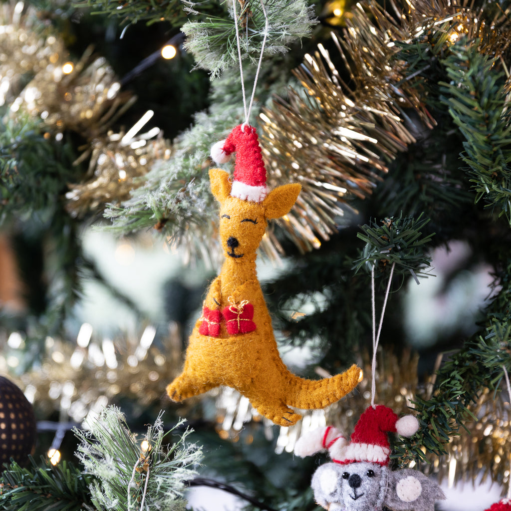 A golden-brown felt kangaroo Christmas ornament wearing a red Santa hat and red gift pouch, hanging on a decorated Christmas tree with lights.