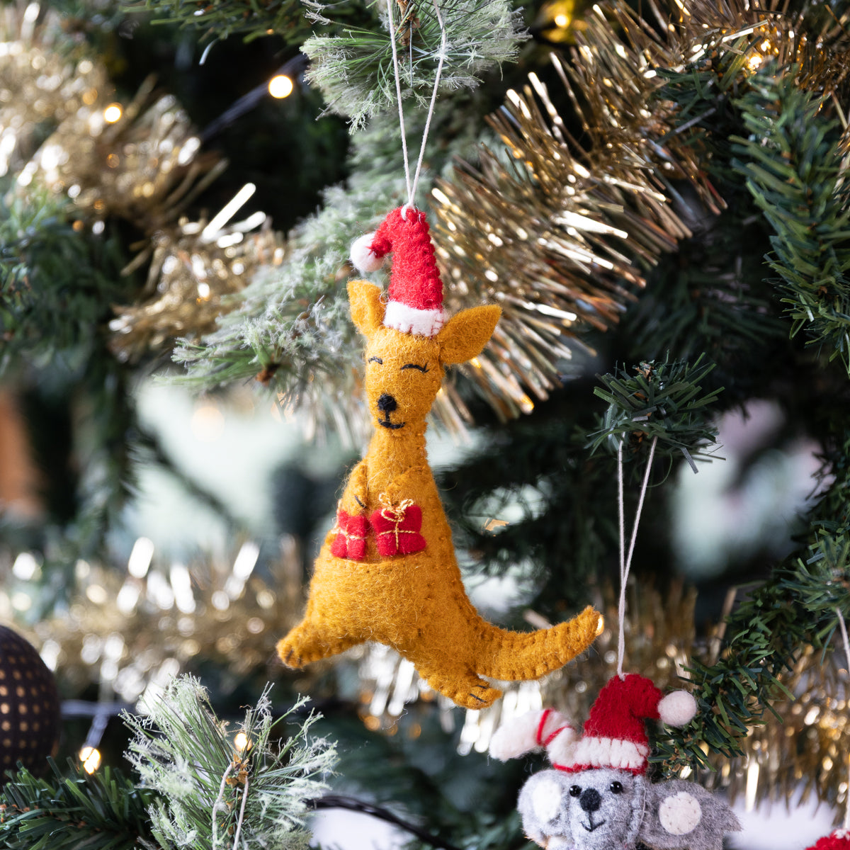 A golden-brown felt kangaroo Christmas ornament wearing a red Santa hat and red gift pouch, hanging on a decorated Christmas tree with lights.