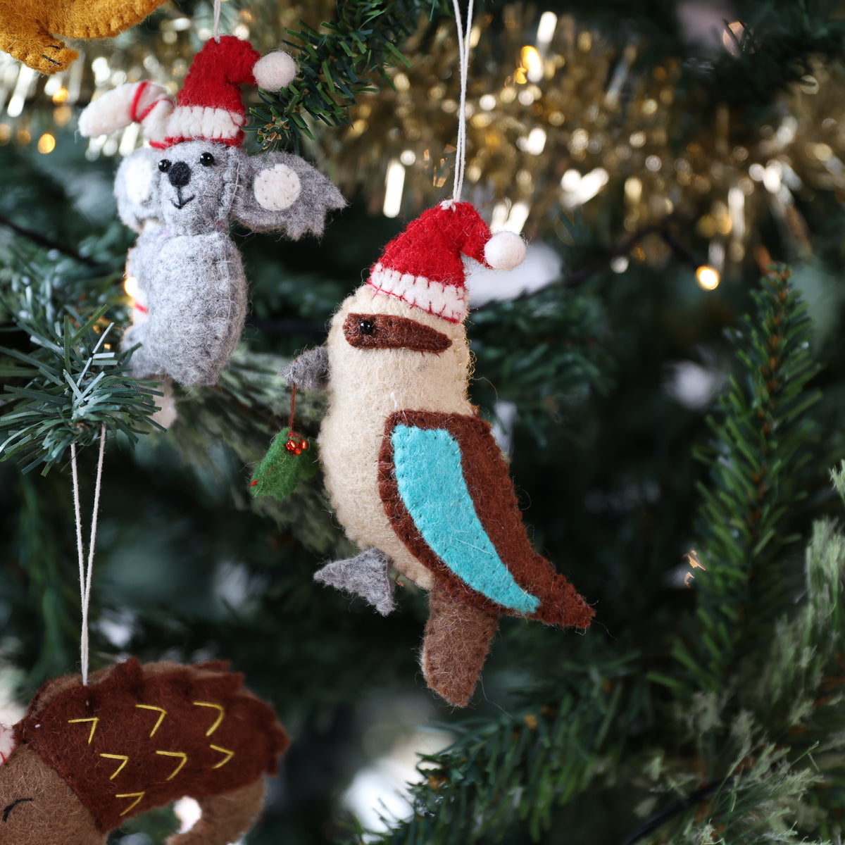 A felt Christmas tree ornament shaped like a brown and cream bird with a turquoise wing and red Santa hat, hanging on a green Christmas tree alongside other animal ornaments.