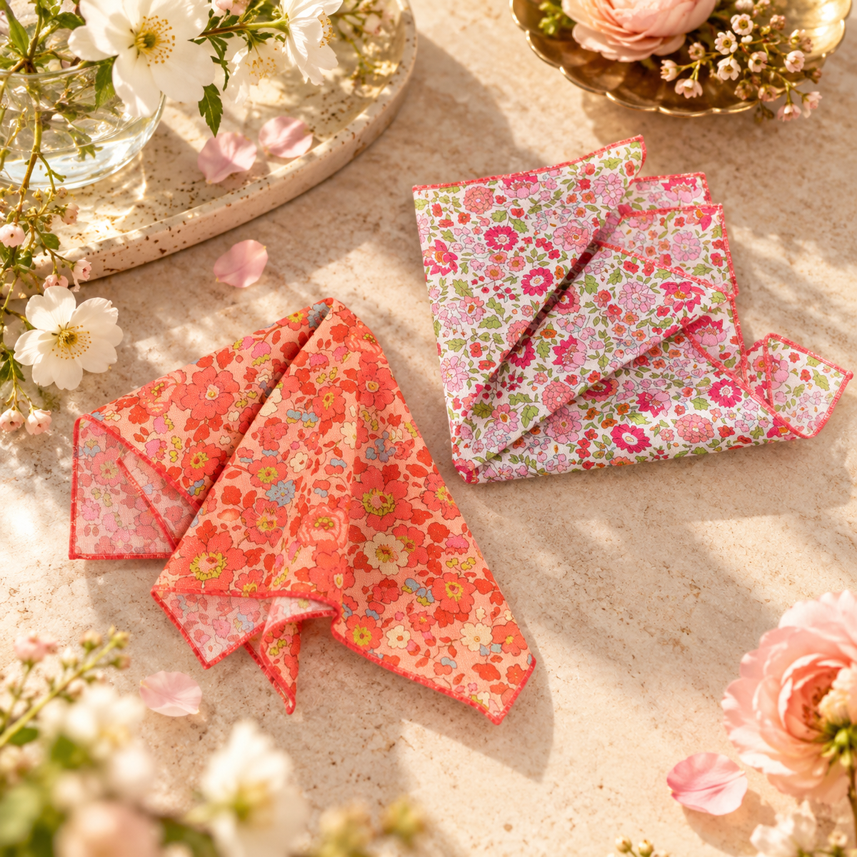 Two folded floral fabric handkerchiefs displayed on a light stone surface with decorative flowers and petals around them. One handkerchief is coral-orange with pink and yellow floral print, and the other is pink with multicolor floral and paisley print.