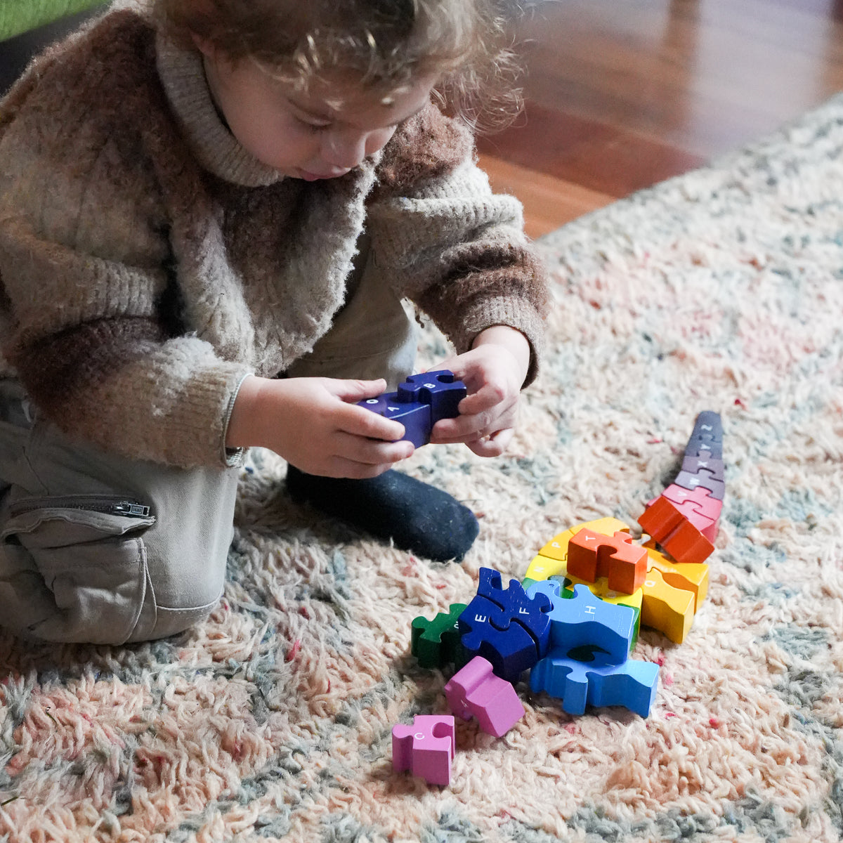 A child in a brown fuzzy sweater kneels on a textured rug, holding a blue wooden block from a colorful set of interlocking toy blocks.