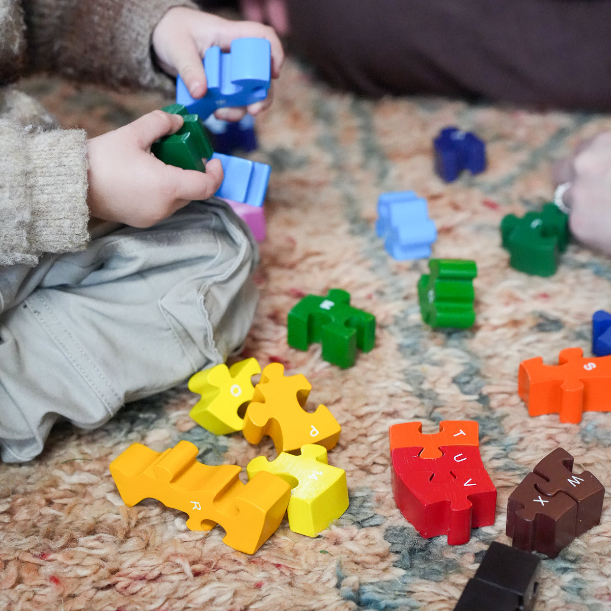 Children playing with colorful alphabet puzzle blocks on a textured carpet. The image shows hands holding blue and green blocks, with yellow, red, orange, brown, and blue pieces scattered around.