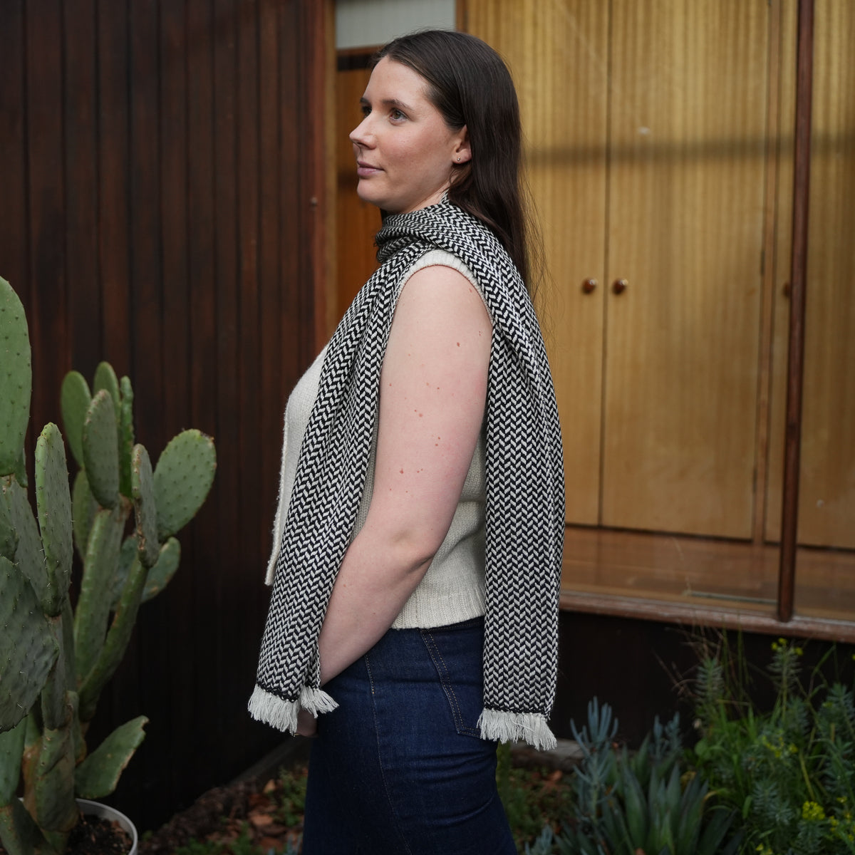 A woman in a side profile wearing a black and white patterned knit scarf with white fringe, styled over a cream sleeveless top and dark blue jeans, standing outdoors in front of a wooden door with plants nearby.