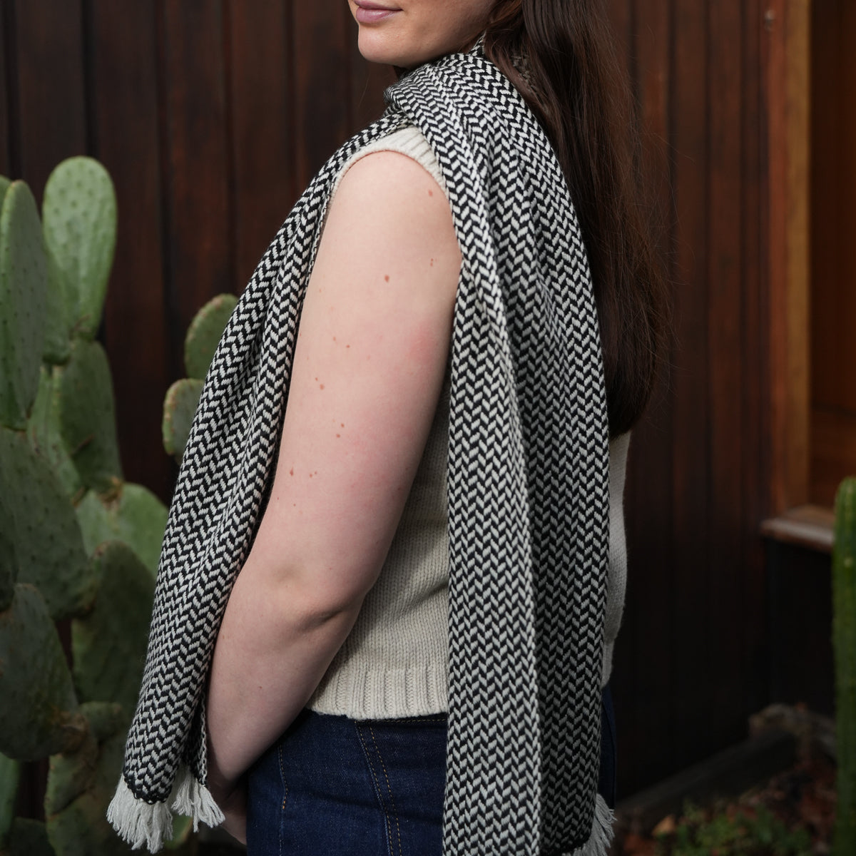 A woman wearing a black and white herringbone patterned knit scarf or shawl draped over her shoulders, paired with a cream sleeveless top and dark blue jeans, standing outdoors in front of a dark wooden fence and cacti.