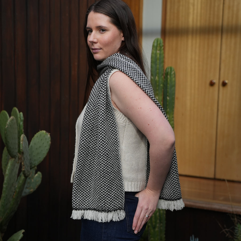 A woman wearing a black and white herringbone patterned knit scarf with white fringe, styled over a cream sleeveless sweater and dark jeans, standing in front of a wooden door and cactus plants.