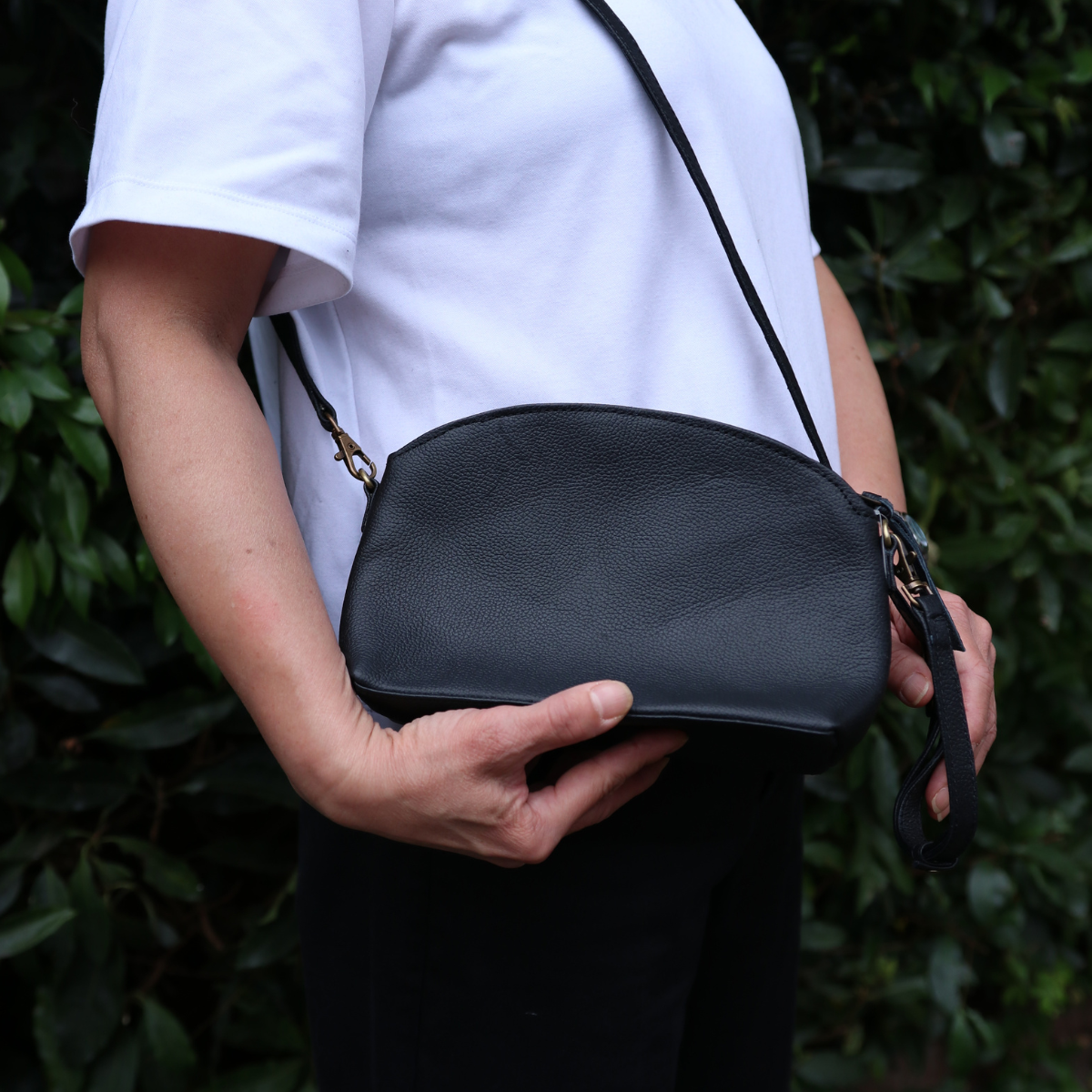Person holding a black leather handbag against a green leafy background