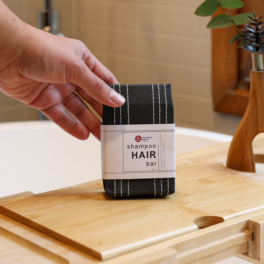A hand holds a packaged shampoo hair bar on a wooden bath tray in a bathroom setting.