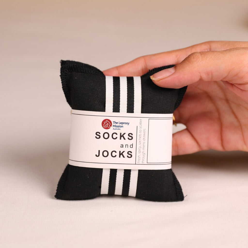 A hand holds a small black fabric sock pillow with white stripes and a white paper label that reads 'The Layneston Mission SOCKS and JOCKS' on a light beige background.