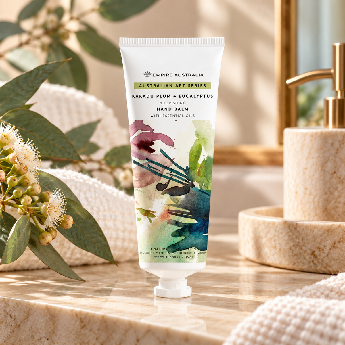 Empire Australia Australian Art Series Karadu Plum + Eucalyptus Hand Balm in a white squeeze tube with a colorful botanical illustration, set on a marble bathroom counter with a soap dispenser and eucalyptus flowers in the background.