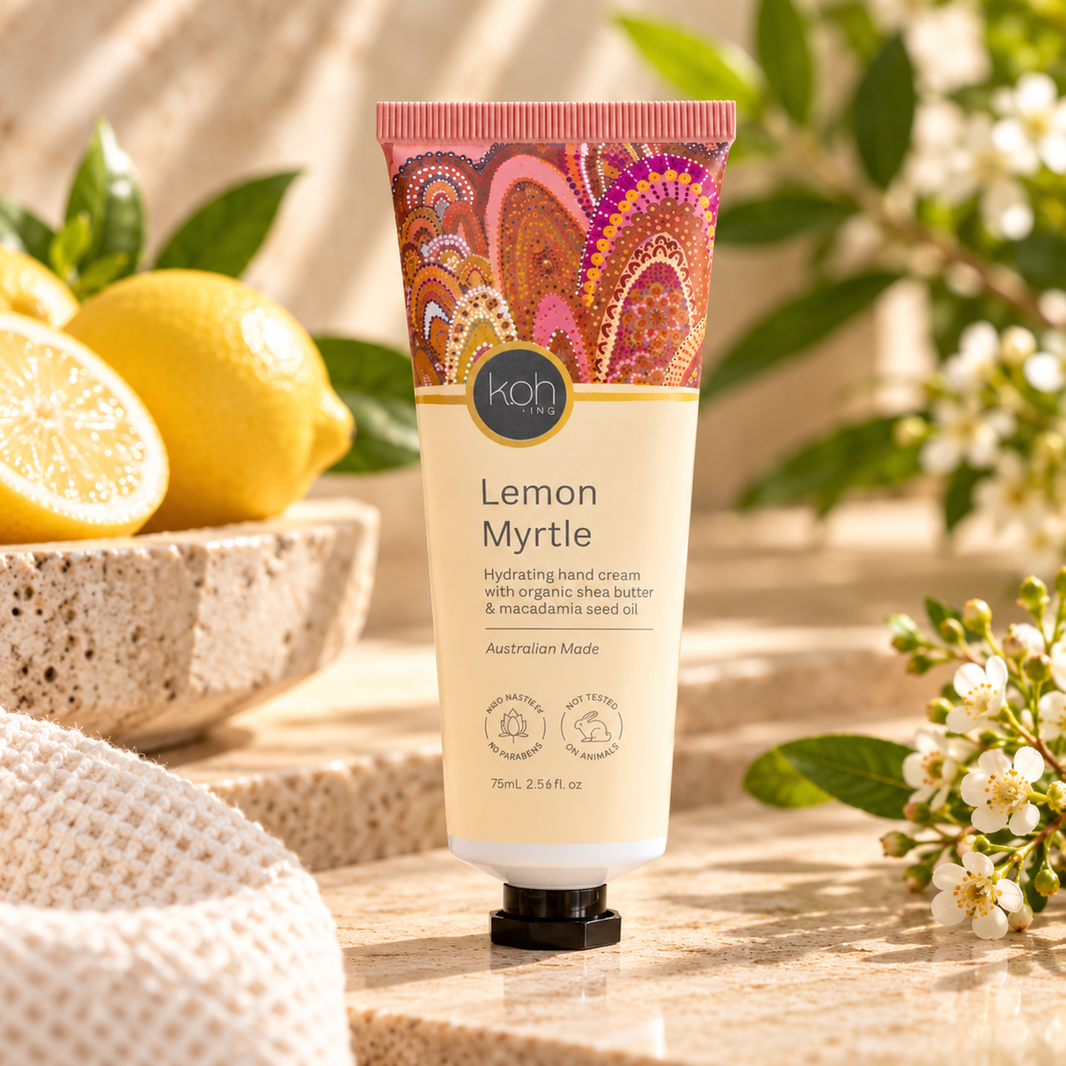 A squeeze tube of Koh Lifestyle Lemon Myrtle hand cream stands on a stone surface with lemons, green leaves, and small white flowers around it. The tube is cream-colored with a pink and orange paisley cap design and a black cap.