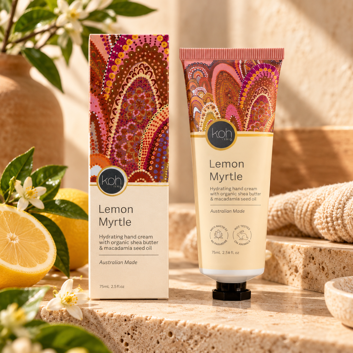 A tube and box of koh Lemon Myrtle hand cream are displayed on a stone surface with lemon slices, flowers, and a sponge in a warm, natural setting.