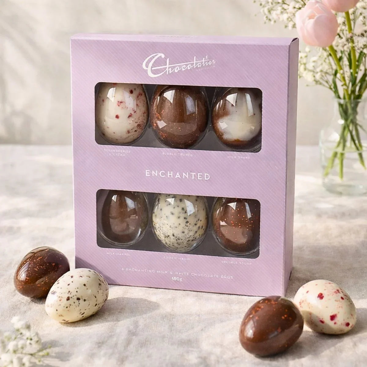 Box of Chocolatier Enchanted chocolate eggs on a light surface with flowers in the background