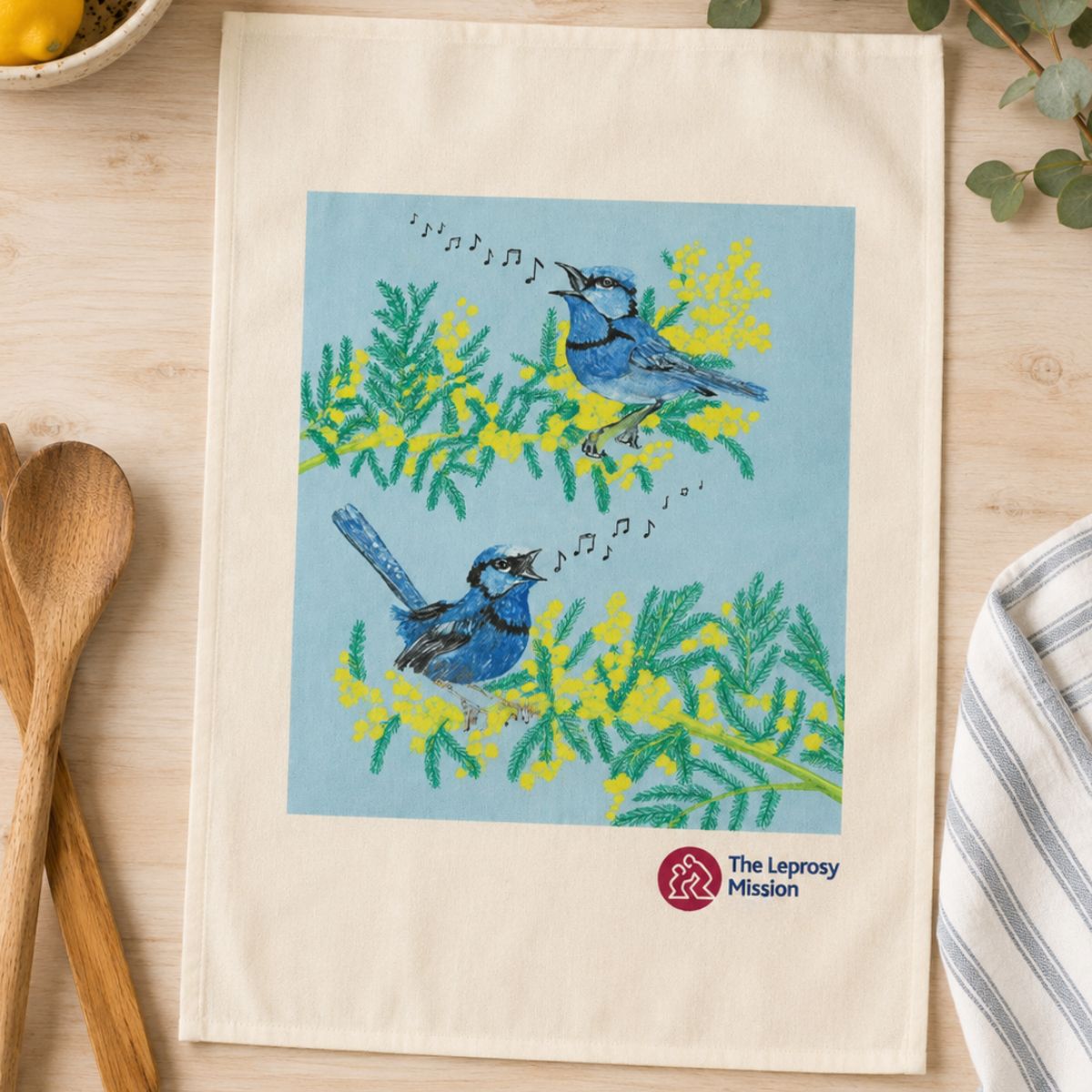 Towel with blue bird design and Leprosy Mission logo on a wooden surface with kitchen items.
