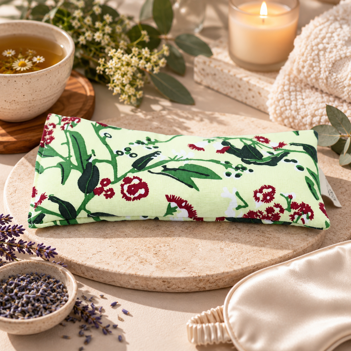 A rectangular eye pillow with a light green floral and bird print rests on a round stone tray in a styled spa setting. Surrounding props include a cup of chamomile tea, lavender, a lit candle, a cream knit blanket, and a satin sleep mask.