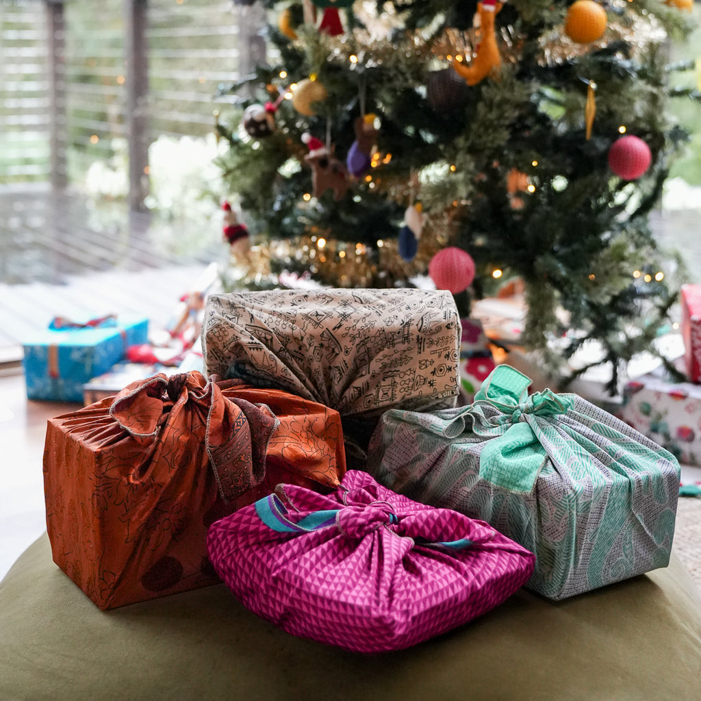 Four wrapped gift packages in colorful patterned fabrics with drawstring bows, arranged on a table in front of a decorated Christmas tree.