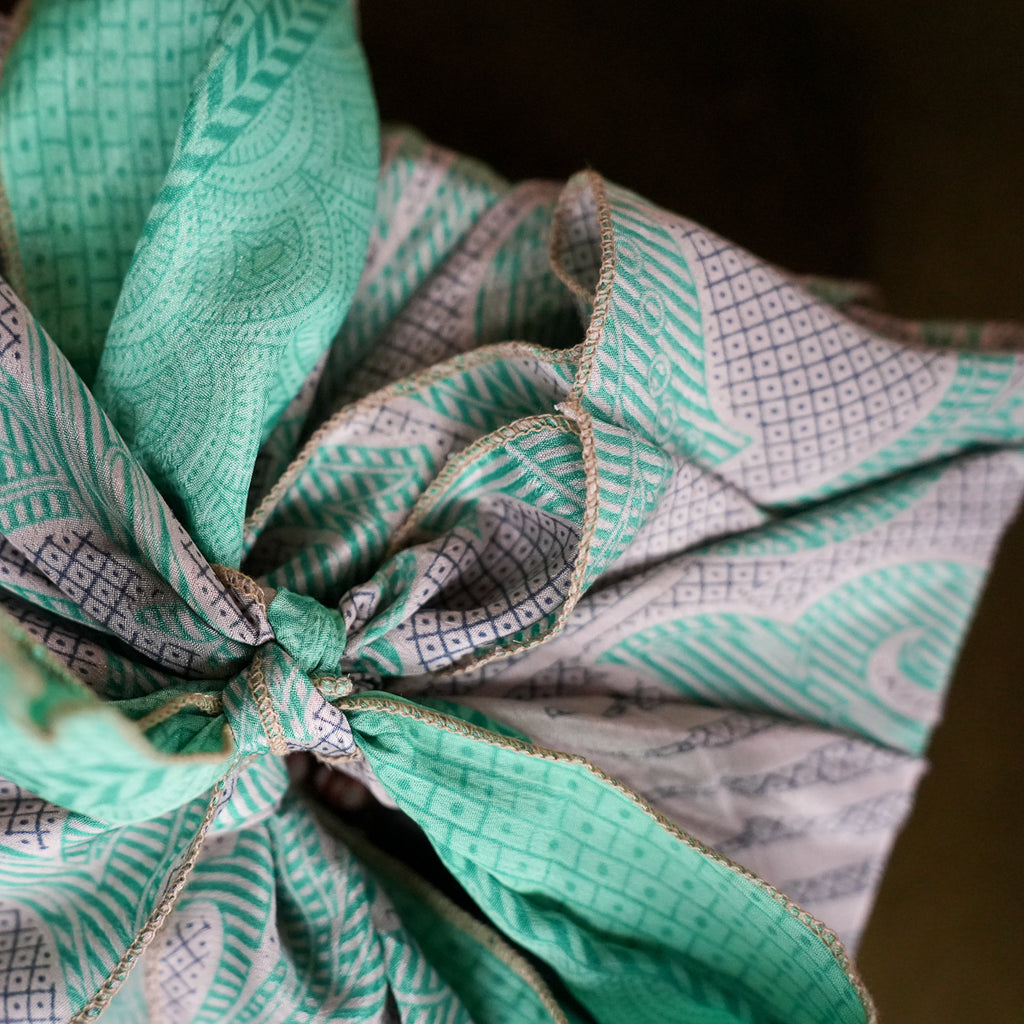 A close-up of a fabric gift wrap tied in a knot, featuring a mint green and white patterned design with a thin beige twine ribbon.