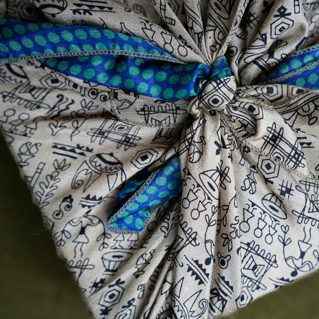 A close-up of a fabric gift wrap tied in a knot, featuring a beige base with black hand-drawn style illustrations and a bright blue and green patterned ribbon accent.