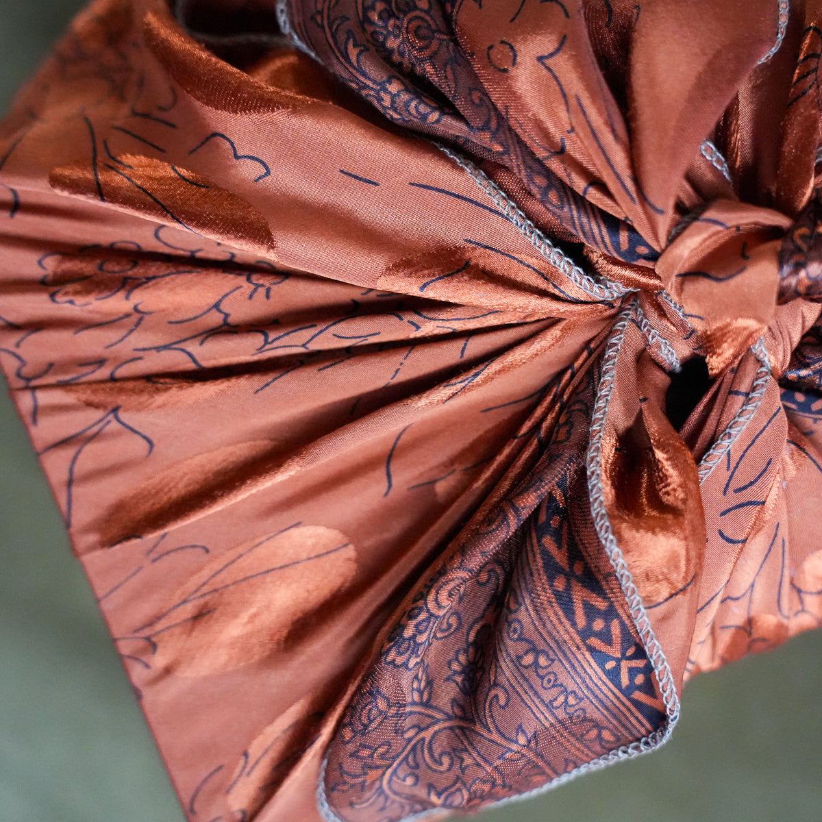 A close-up of a gathered, metallic copper-brown fabric or scarf with dark blue abstract line patterns and a patterned reverse side, shown against a muted green-gray background.
