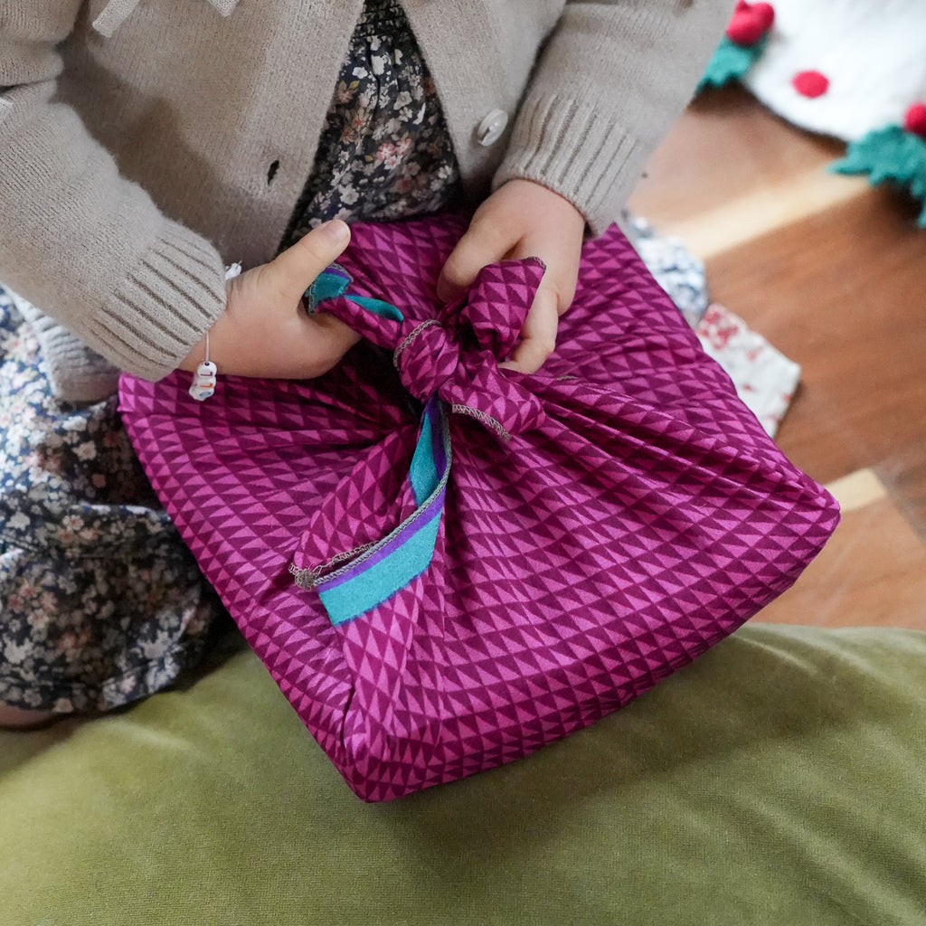 A close-up of a person holding a wrapped gift package in a traditional Japanese style, featuring a bright magenta geometric pattern and a turquoise ribbon bow, set against a cozy indoor background.