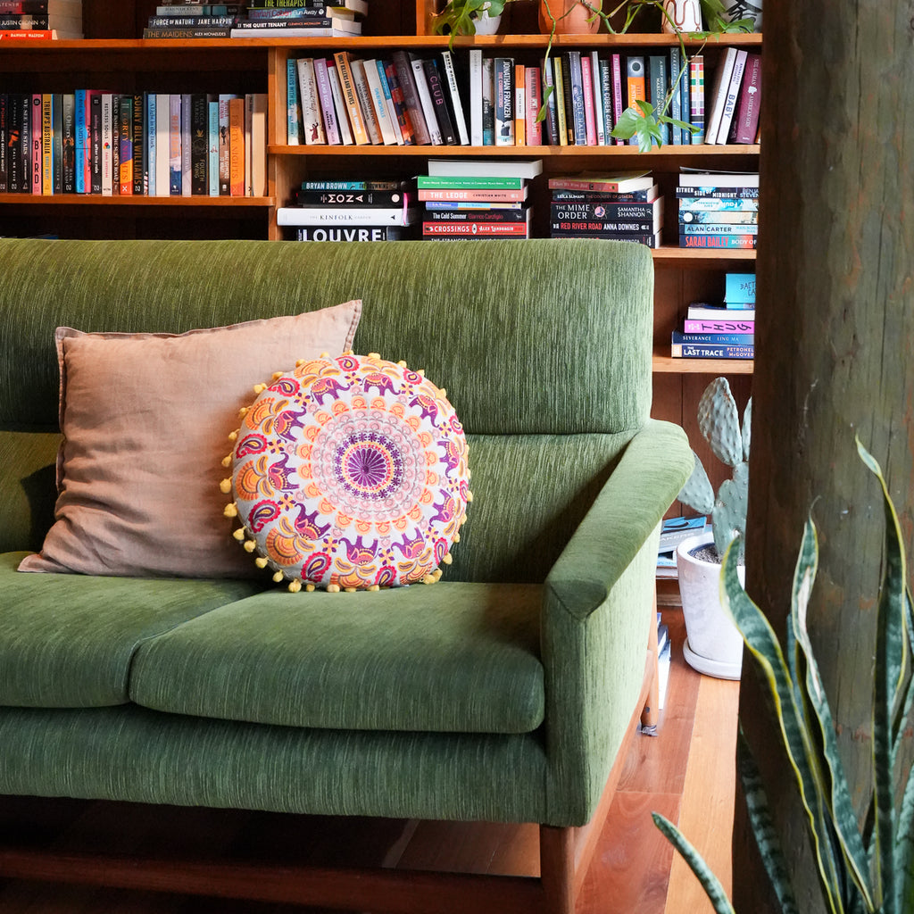 An olive green upholstered loveseat in a cozy room setting, styled with a beige throw pillow and a round, colorful mandala-patterned decorative pillow with pom-poms. Bookshelves and houseplants are visible in the background.