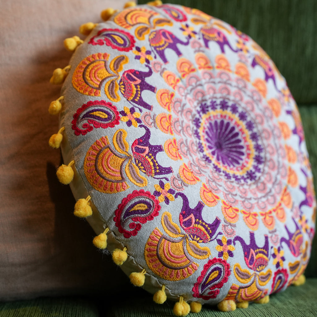 A round decorative throw pillow with a colorful embroidered mandala and elephant pattern, finished with yellow pom-pom trim, resting against a neutral surface.