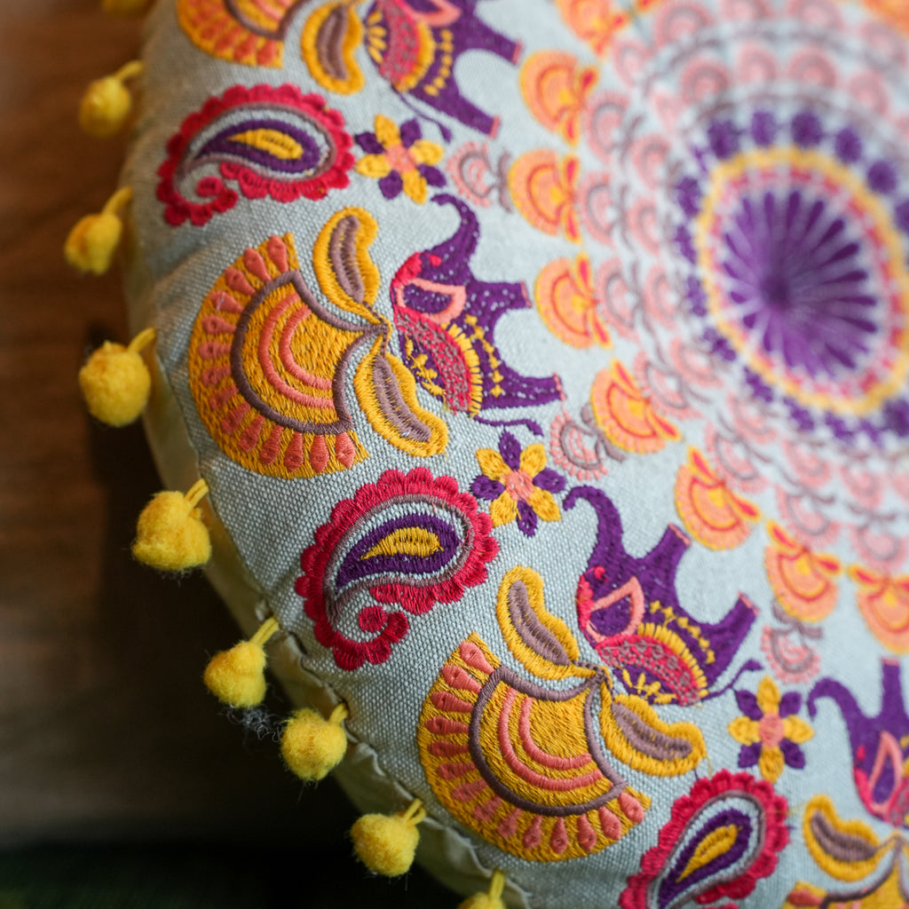 Close-up of a decorative cushion cover with a colorful embroidered paisley and mandala pattern in purple, orange, yellow, and red on a light gray fabric, finished with a row of yellow pom-poms along the edge.