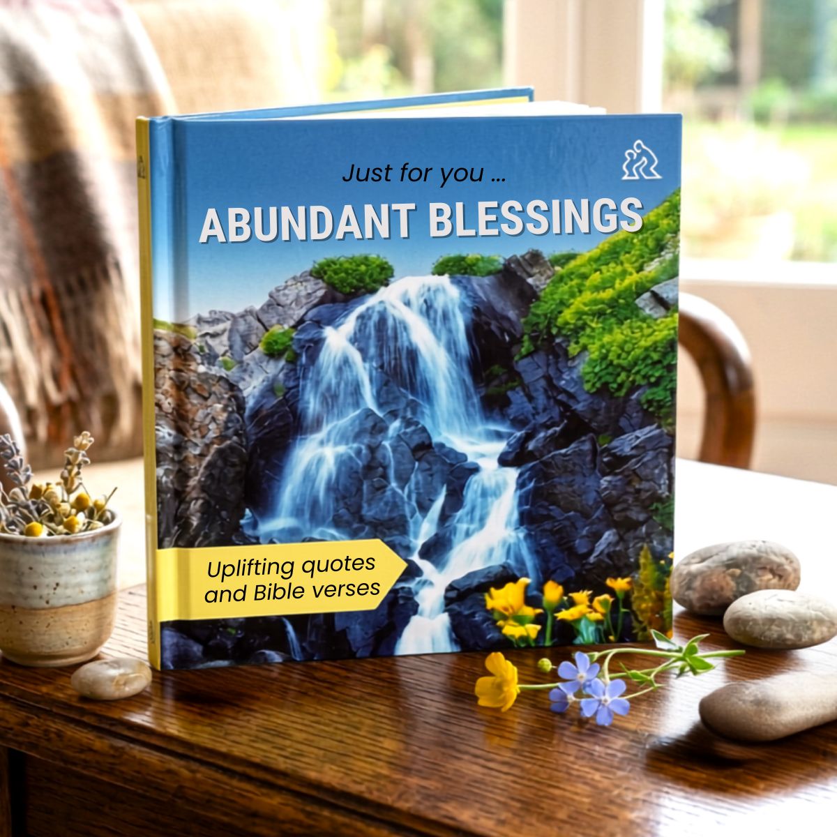 Book titled 'Abundant Blessings' with a waterfall image on a wooden surface with flowers and stones.