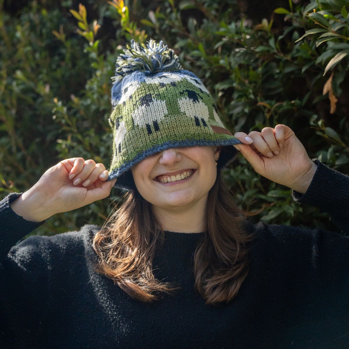 Person wearing a green knitted hat with a pattern, holding it over their eyes, with a blurred natural background.