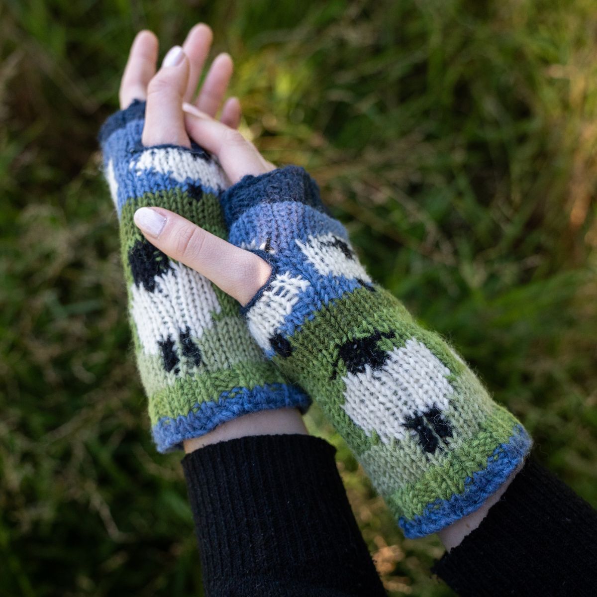 Knitted fingerless gloves with a pattern of black shapes on a green and blue background, held against a grassy background.