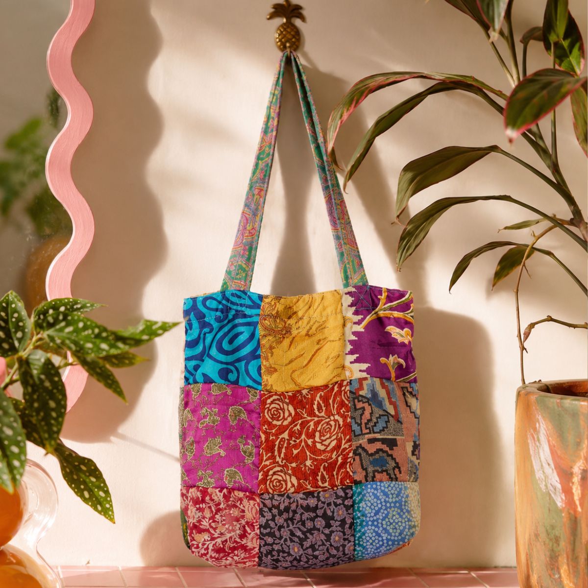 Colorful patchwork tote bag with floral patterns hanging against a neutral wall with plants.