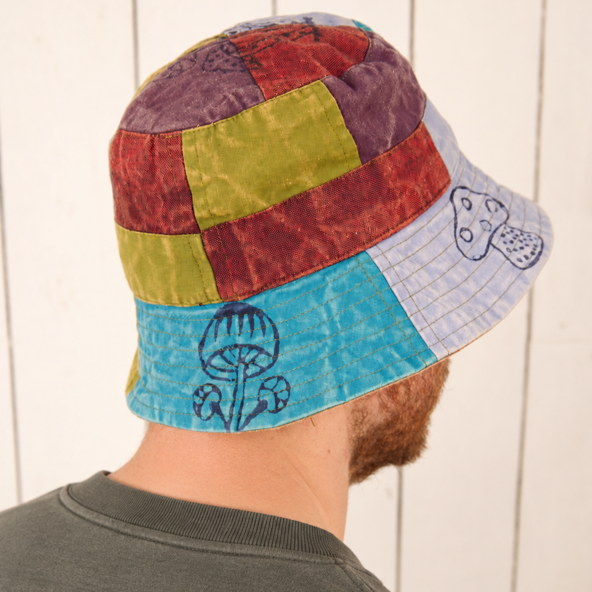 A man wearing a colorful patchwork bucket hat with mushroom illustrations, photographed from the back against a white wood-paneled wall.