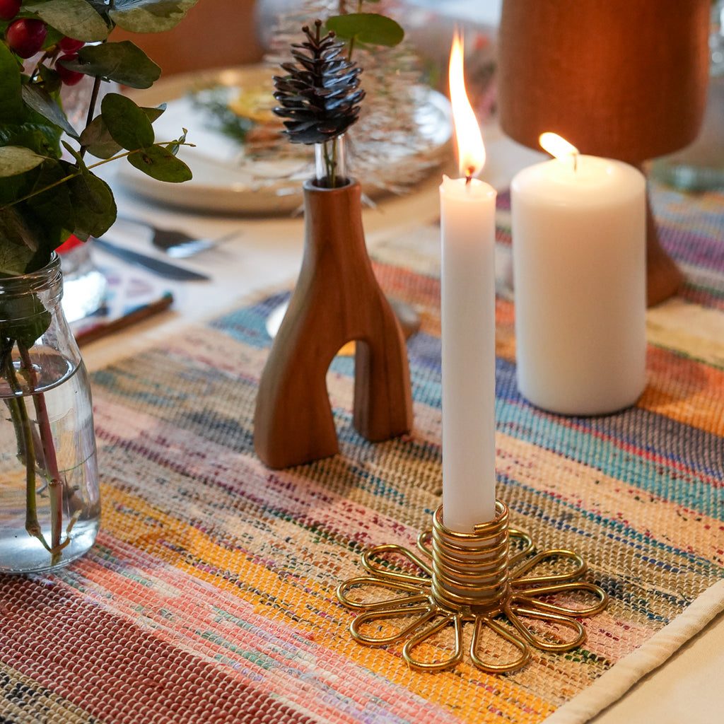 A brass flower-shaped candle holder with a lit white taper candle, set on a colorful woven table runner alongside other home decor items including a wooden vase with a pinecone and a white pillar candle.