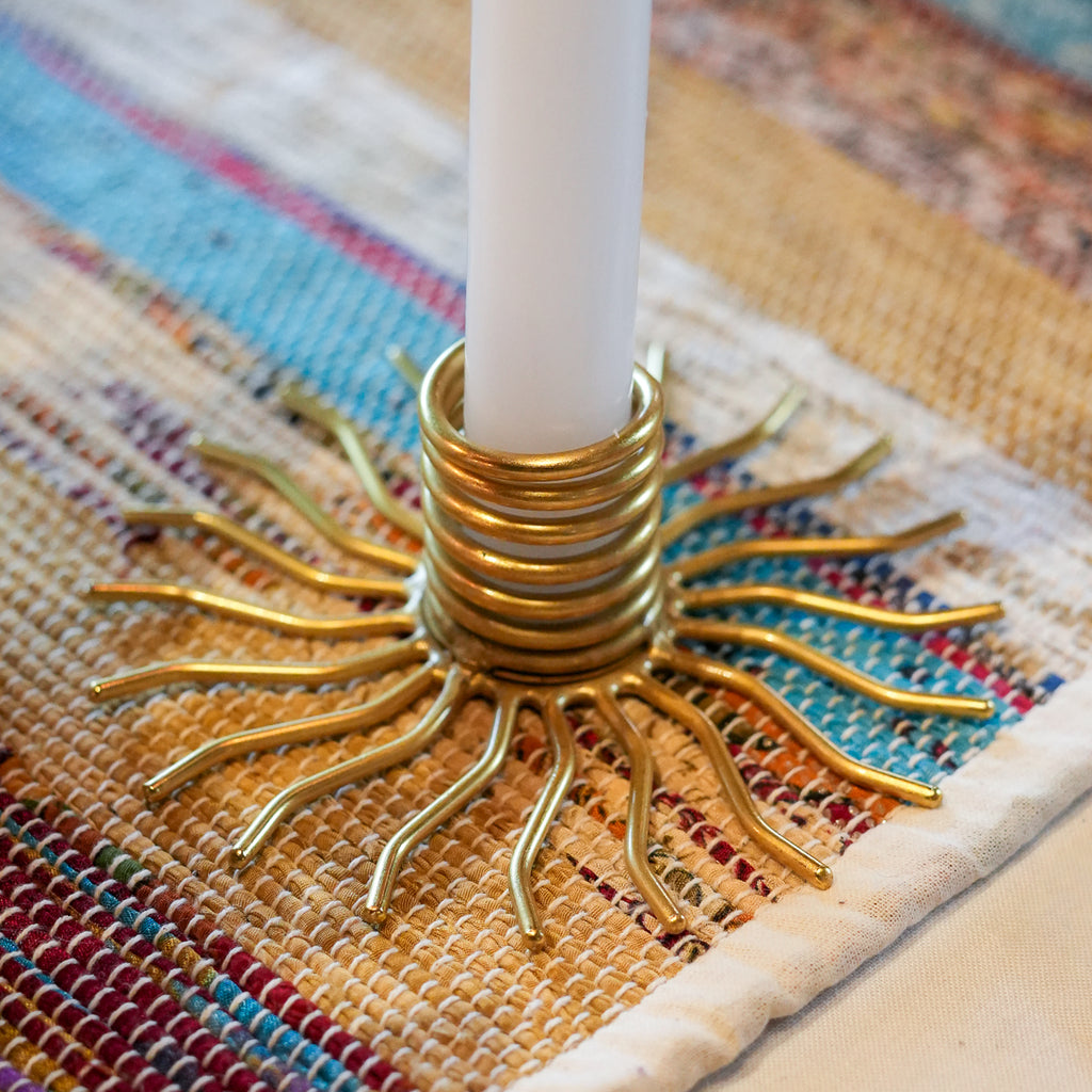 A close-up of a gold metal sunburst candle holder with a white candle inserted, resting on a woven multicolor table runner.