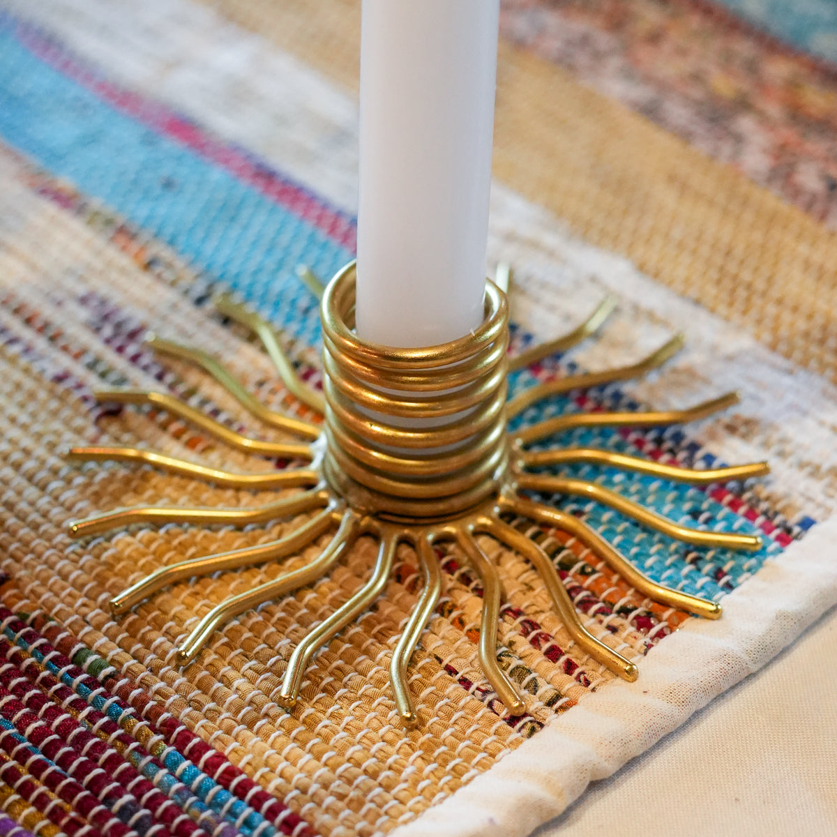 A close-up of a gold metal sunburst candle holder with a white candle inserted, resting on a woven multicolor table runner.