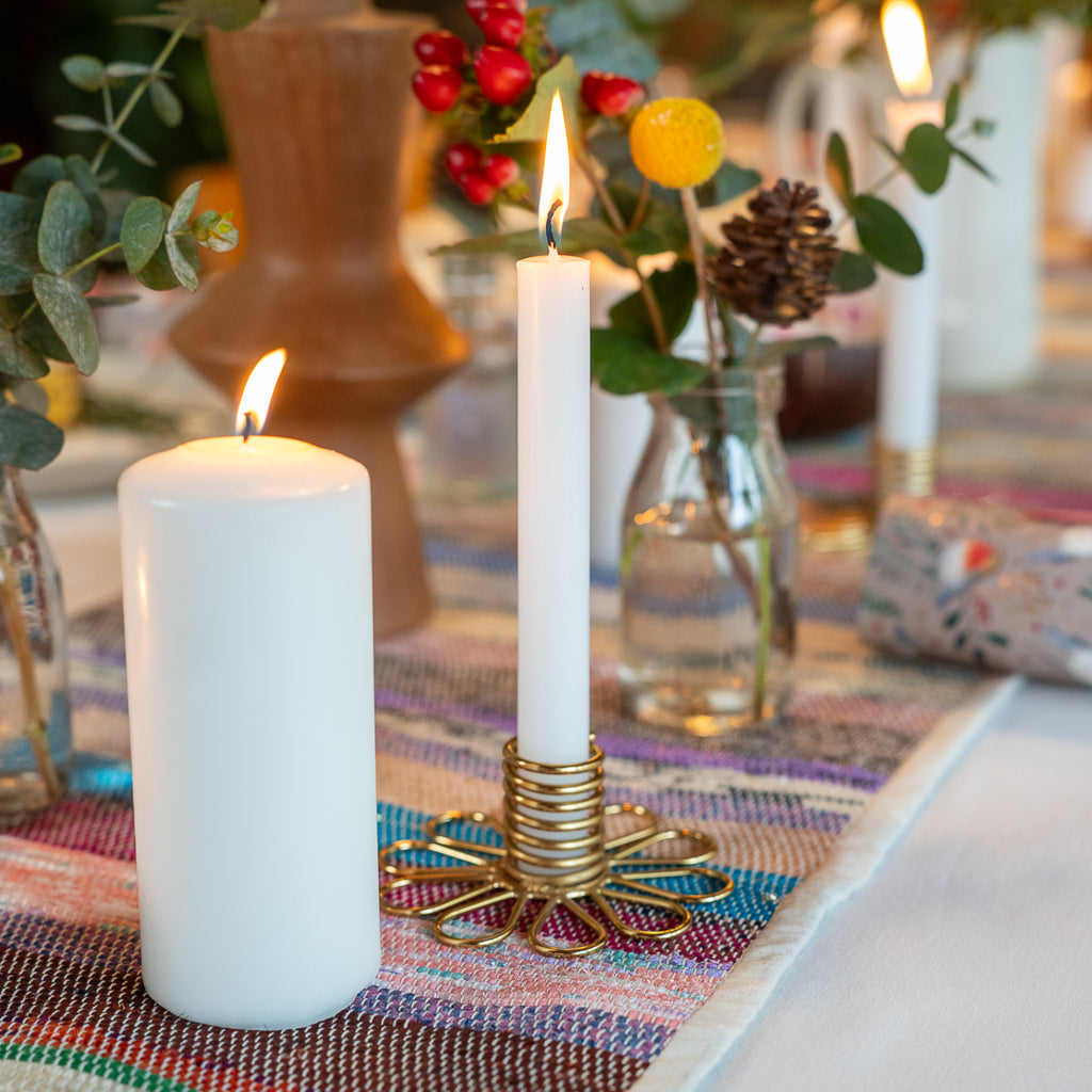 A lit white pillar candle in a gold spiral holder on a woven multicolor table runner, with other candles and floral arrangements blurred in the background.