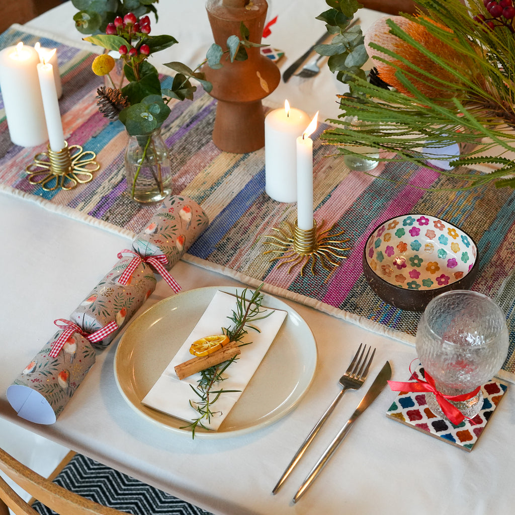 A styled holiday table setting with a colorful striped runner, lit white pillar candles, a patterned ceramic bowl, a wrapped gift tied with a red gingham ribbon, silver cutlery, and a clear textured glass on a white tablecloth.
